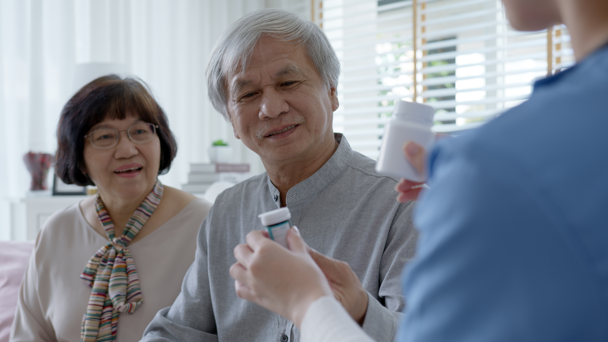 Young caregiver in scrubs uniform showing medicine bottle to elderly asian couple man and woman in home visit care nursing service. Asian senior with assisted living medication monitoring concept.